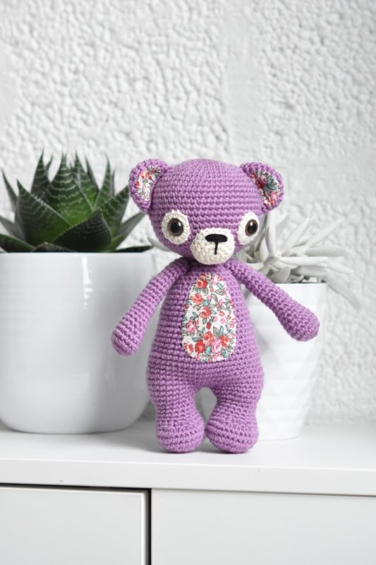 Purple crochet teddy bear standing in front of the plants